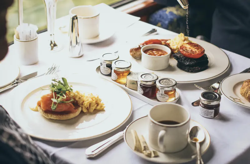 Luxury train dining table with smoked salmon eggs Benedict, black pudding, tea, coffee, jams, and window view.