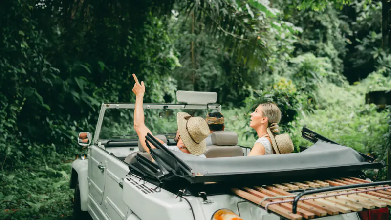 Couple in white convertible Jeep, man pointing up, woman smiling, driving through lush Bali jungle at Buahan.