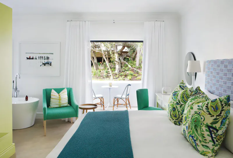 Elegant hotel bedroom with teal bedspread, green pillows, freestanding tub, and window view of lush greenery
