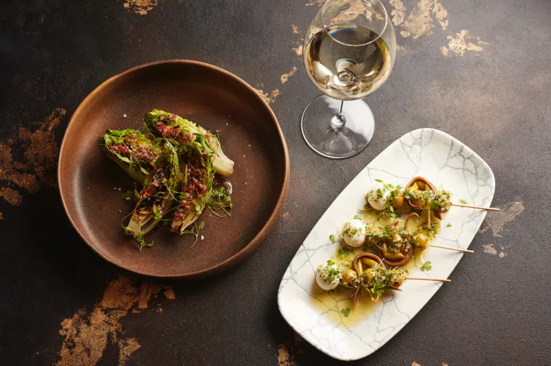 El Norte dishes: grilled endive in wooden bowl and white cheese skewers on marble plate with white wine glass