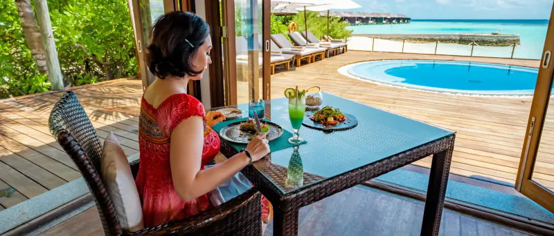 Woman in red dress eating at glass table with green juice and fruits, overlooking ocean and infinity pool at tropical resort