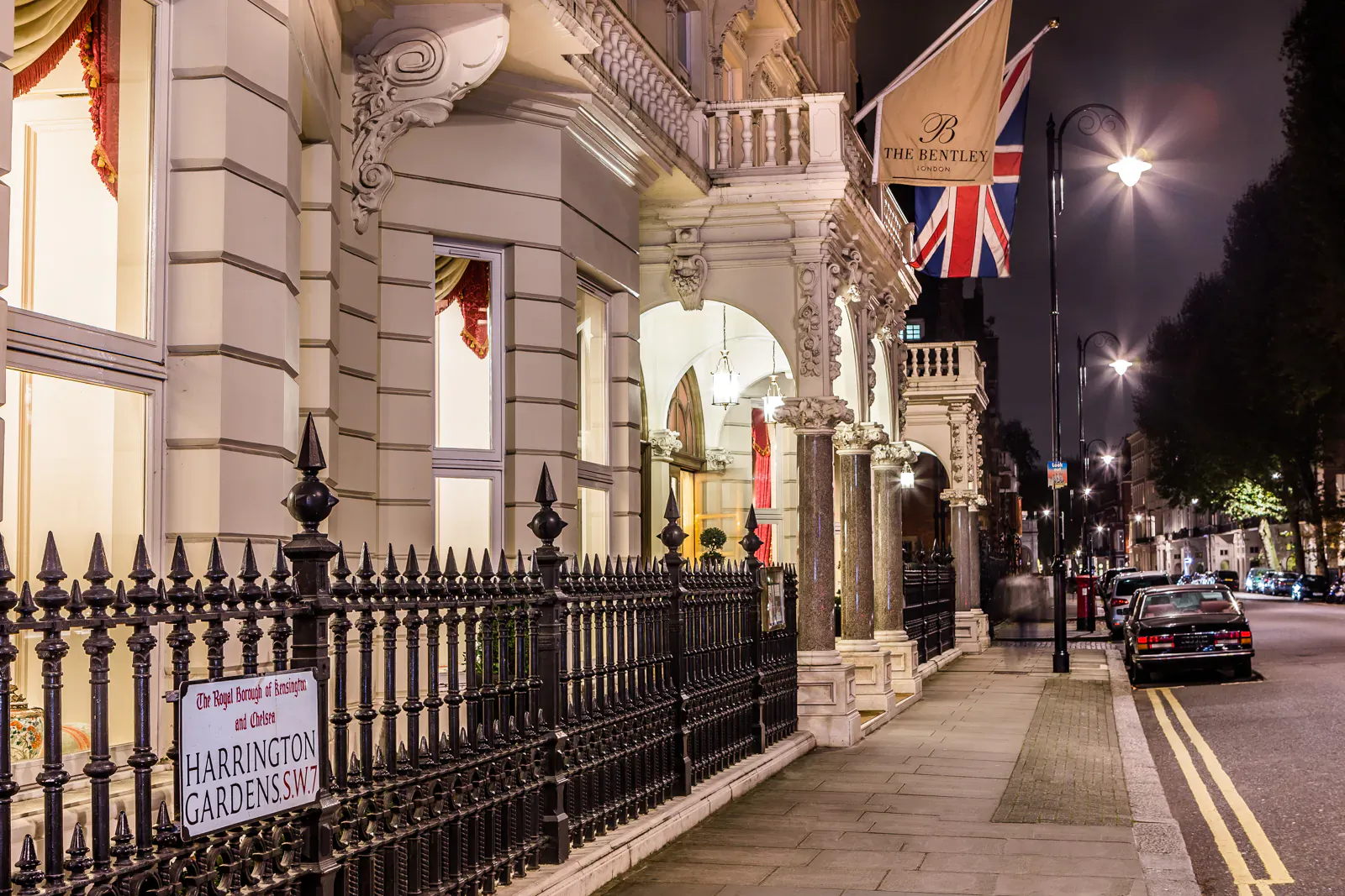 Bentley Hotel facade in Kensington, London at night, with Union Jack flag, 'Harrington Gardens' sign, and streetlights.