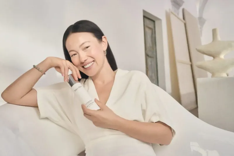 Smiling Asian woman in white dress holds white skincare bottle, seated in modern art-filled room