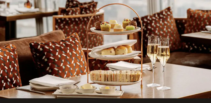 Three-tiered stand with scones, pastries, clotted cream, and two champagne flutes on a table in a luxurious London setting.