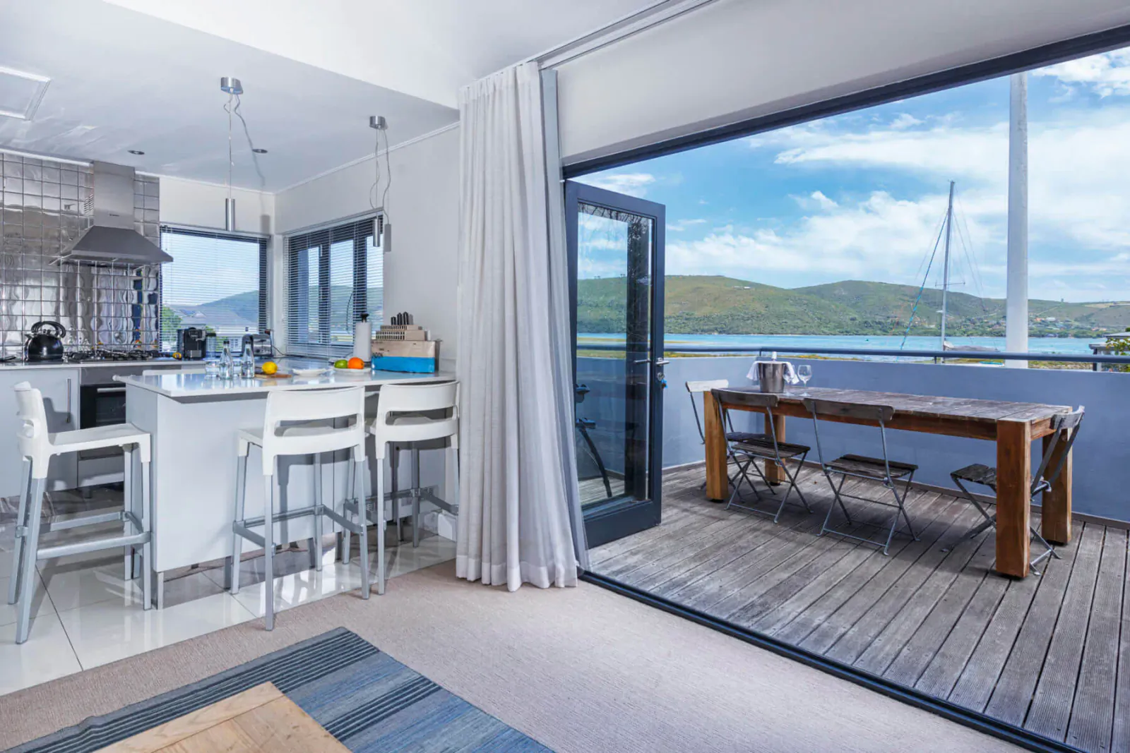 Modern kitchen in Turbine Hotel with white bar stools, wooden dining table, open doors to deck overlooking lake and mountains.