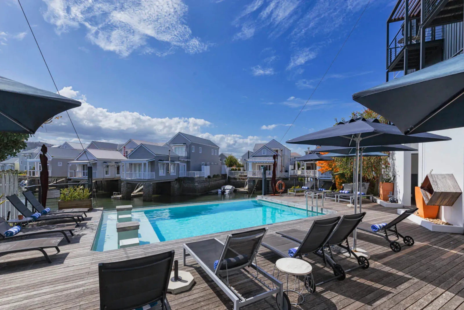 Infinity pool on wooden deck at Turbine Hotel, with lounge chairs, umbrellas, white buildings, and canal under blue sky