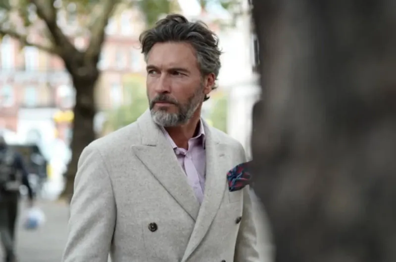 Man with gray beard in light beige double-breasted suit, pink shirt, red pocket square, stands by tree on urban street