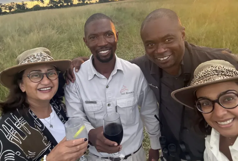 Four smiling people in safari hats and uniforms holding drinks, posing in golden grassy field at sunset