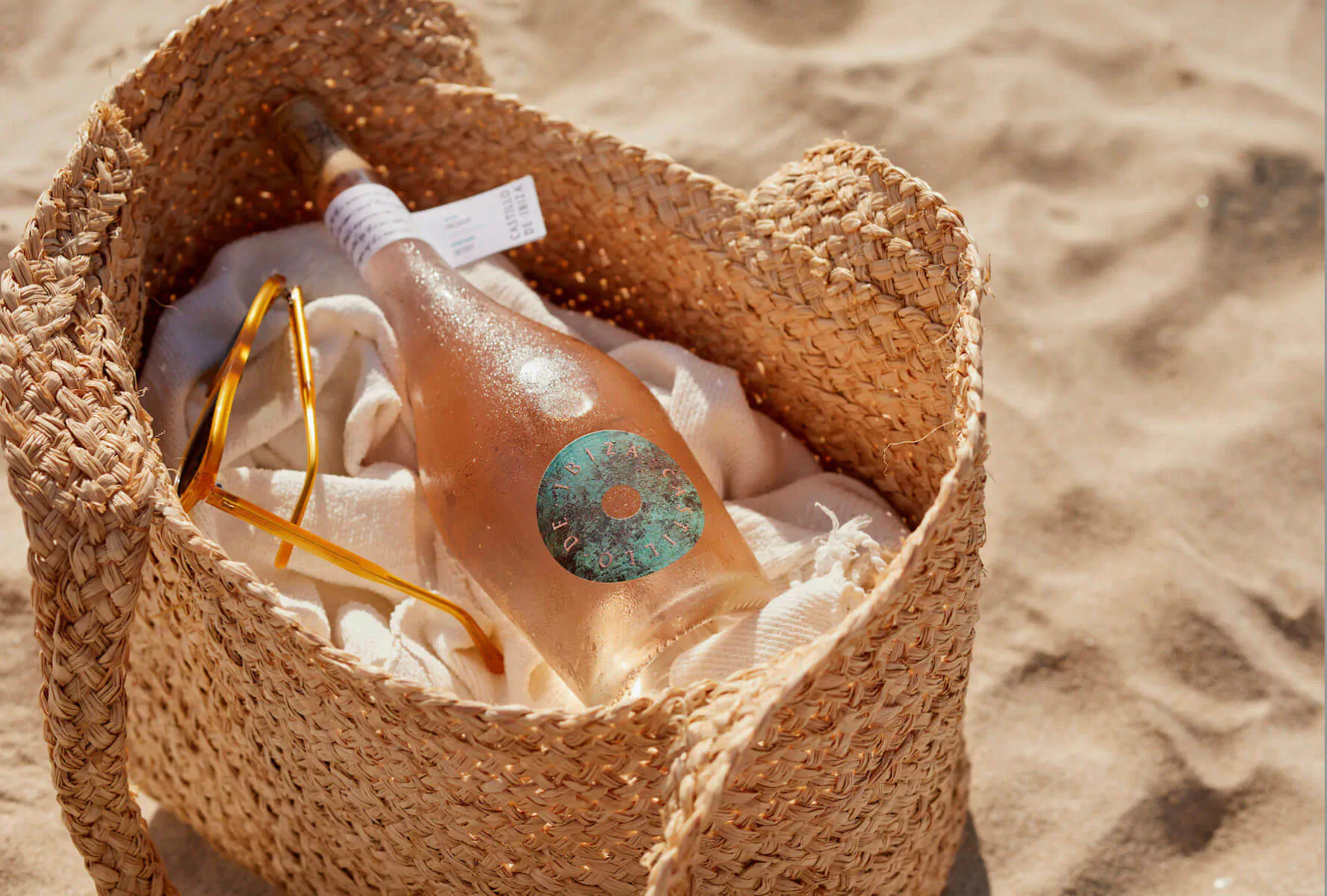 Rosé wine bottle with sunset-inspired label in straw beach bag on sand, with sunglasses