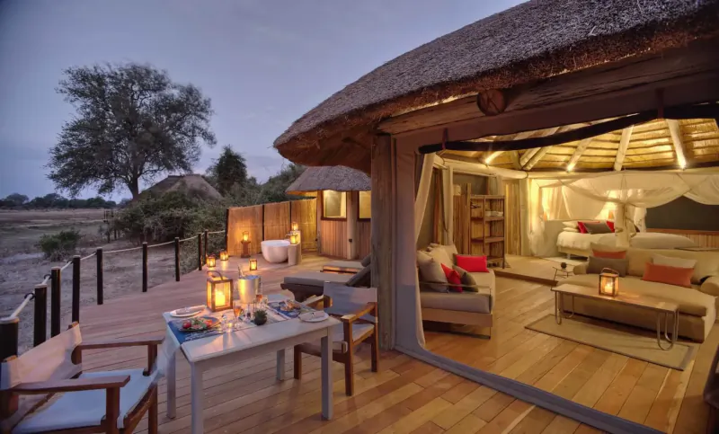 Luxurious Lion Camp tent at dusk with thatched roof, open deck dining table with lanterns, cozy bedroom interior, savanna backdrop.