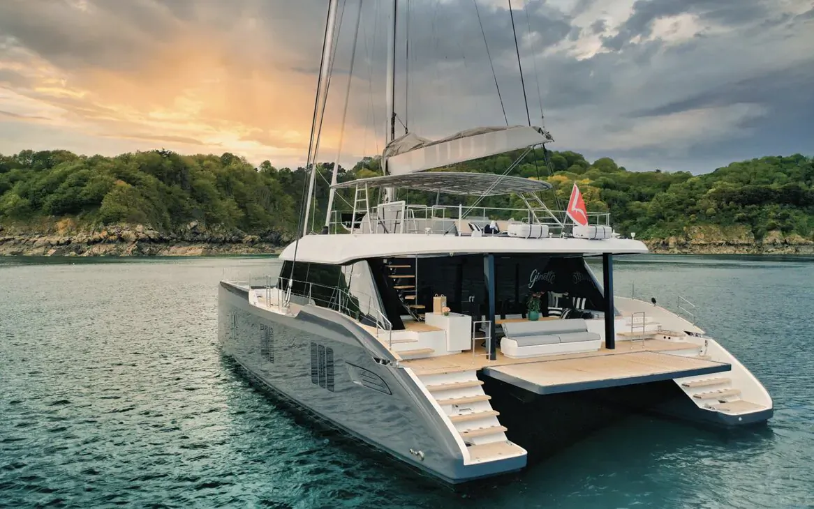 Sleek white Sunreef catamaran yacht docked on turquoise water at sunset, surrounded by lush green hills and cloudy sky.