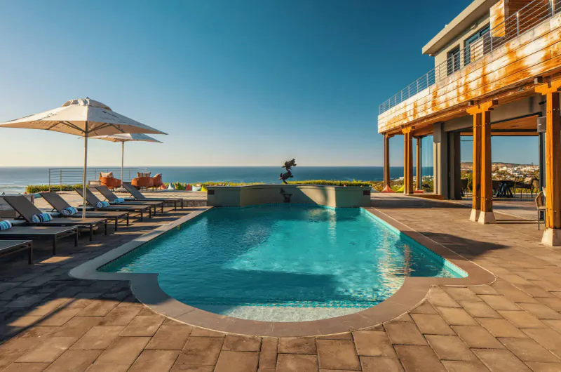Infinity pool at Sky Villa Boutique Hotel overlooking ocean, with lounge chairs, umbrellas, and modern wooden house under blue sky.