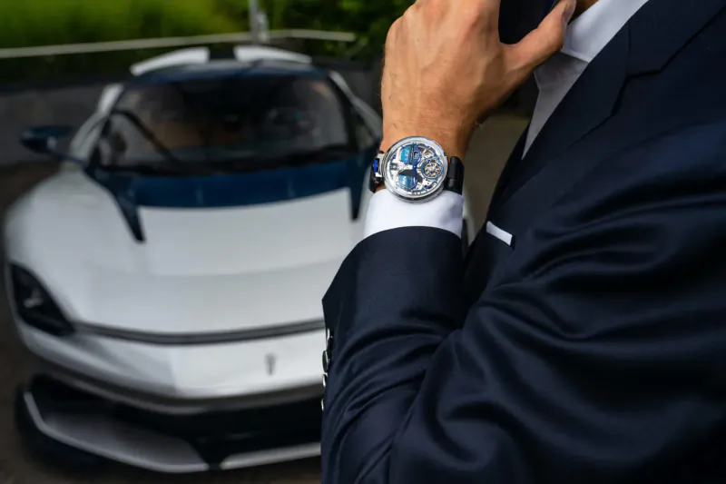 Man in navy suit showing blue BOVET Battista Tourbillon watch beside white luxury sports car