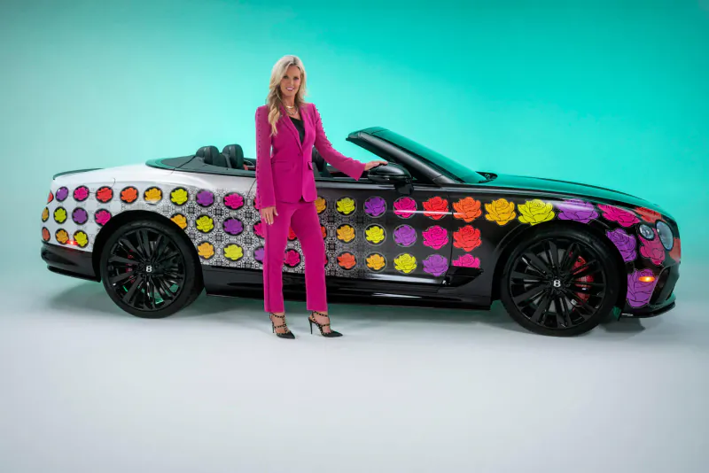 Blonde woman in pink suit poses beside vibrant polka-dot Bentley convertible on turquoise background
