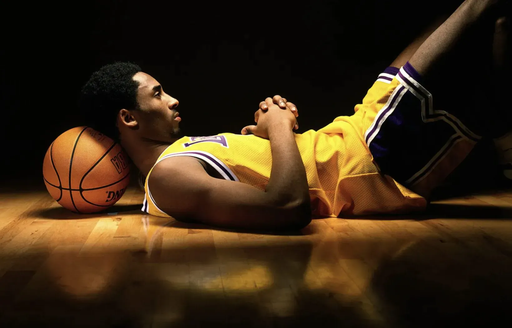 Kobe Bryant lying on basketball court, hands clasped, resting head on basketball, in Lakers jersey and shorts