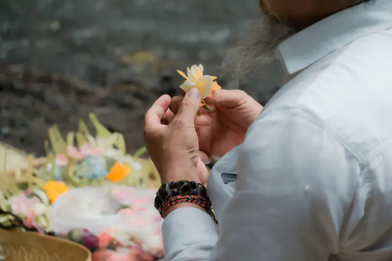Man with long beard holding white frangipani flower amid colorful Balinese offerings at wellness retreat