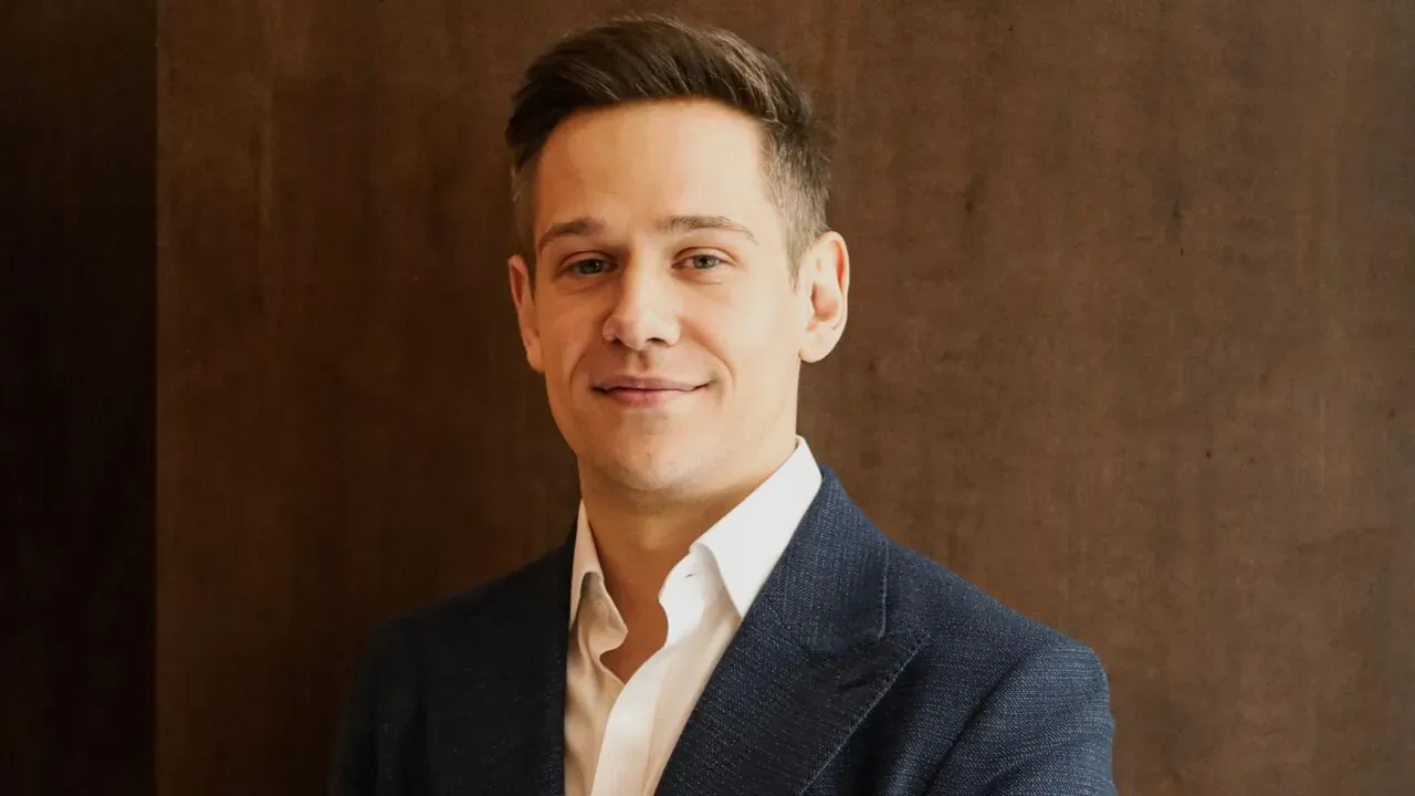 Dr Adam Nusenbaum in navy suit and white shirt, smiling against wood-paneled background