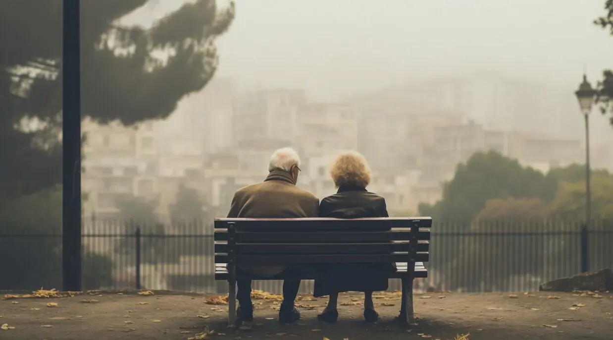 Elderly white-haired couple sits back-to-back on park bench overlooking misty cityscape and trees.
