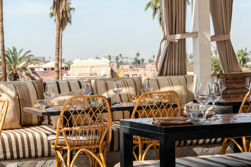 Rooftop restaurant in Marrakech with rattan chairs, beige cushions, wooden tables set with wine glasses, palm trees, and city skyline.