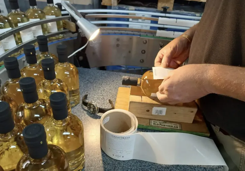 Worker labeling whisky bottles on production line, GreatDrams facility, with conveyor and wooden box.