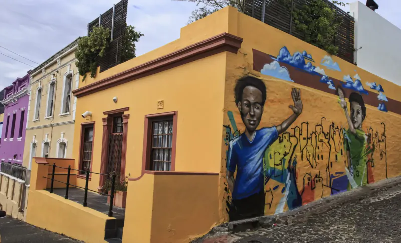 Vibrant street art of young Black boys in blue and green on yellow Bo-Kaap house amid colorful buildings