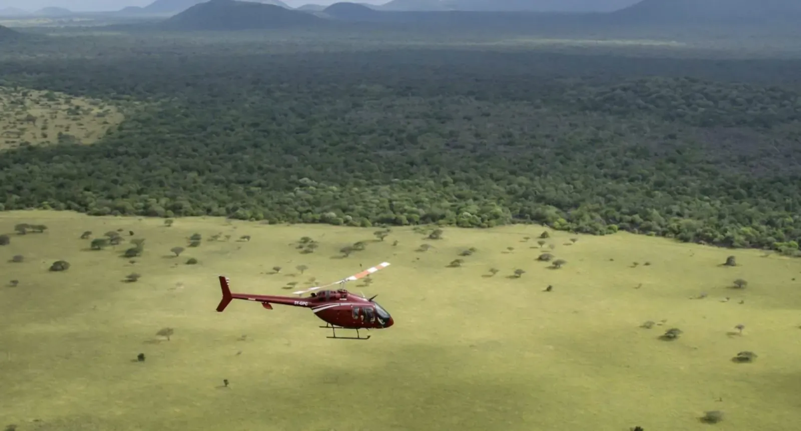 Red Bell 505 helicopter flying over green savanna plains and forested mountains in Kenya