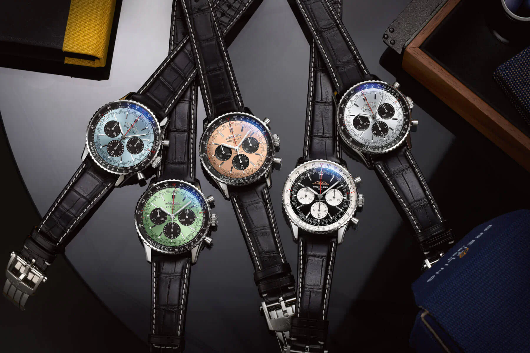 Five colorful Breitling Navitimer chronograph watches on black leather straps, displayed on dark surface with leather folders.