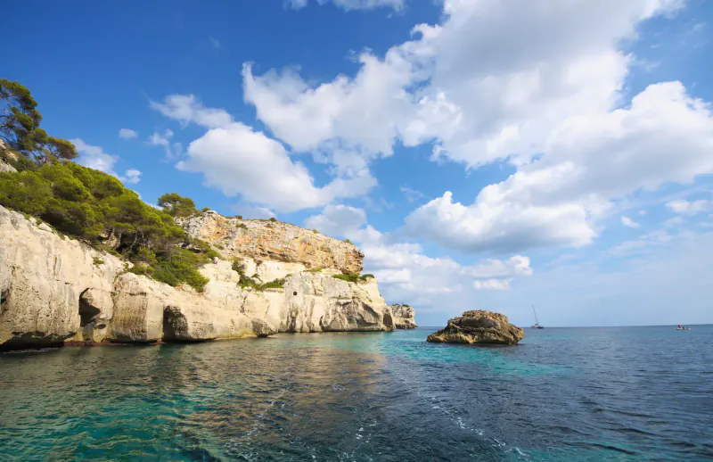 Turquoise sea laps rocky cliffs with pine trees under partly cloudy blue sky, Mediterranean cove