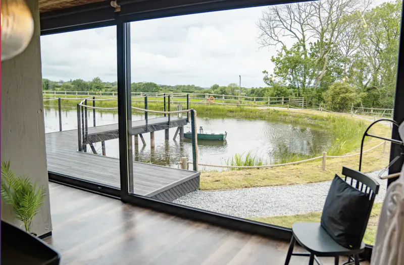 Inside The Boathouse luxury lodge, panoramic window view of pond with green boat at wooden pier, rural fields, chairs nearby.