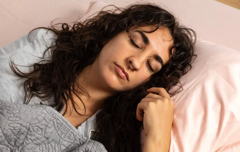 Woman with curly brown hair sleeping peacefully on pink pillow, grey quilt covering her in bed