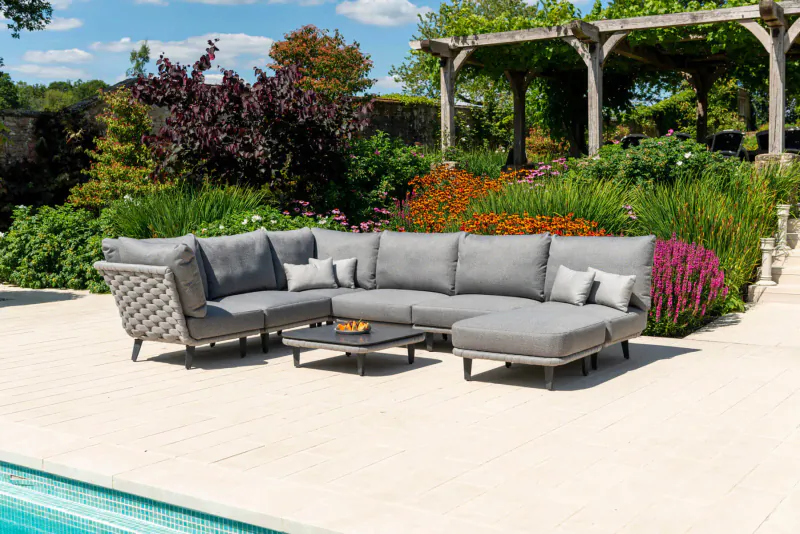 Grey Alexander Rose L-shaped outdoor sofa set with coffee table on patio by pool, surrounded by lush garden and pergola.
