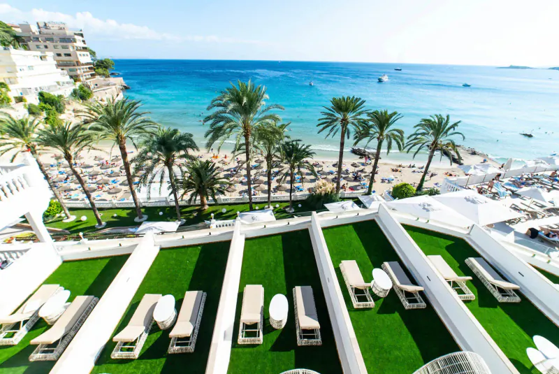 Aerial view of Nixe Palace's white lounge chairs on green terrace overlooking palm-fringed beach and sea.