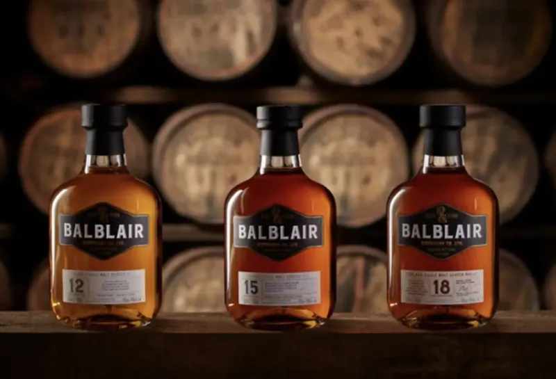 Three Balblair whisky bottles (12, 15, 16 years) on wooden table with oak barrels in background.