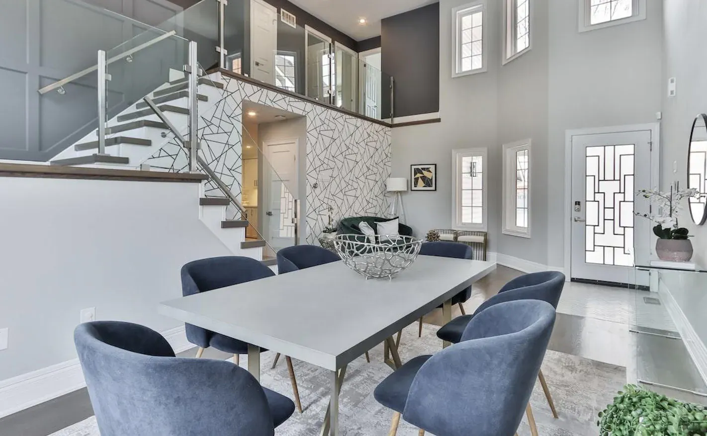 Modern luxury dining room with white table, blue velvet chairs, glass staircase, and elegant decor