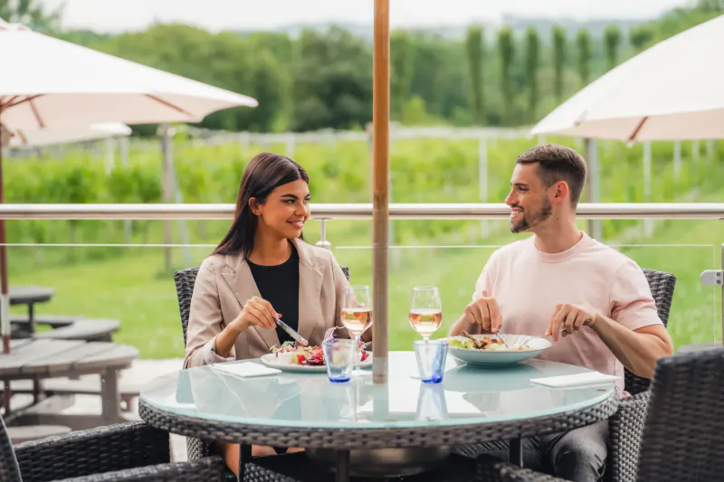 Smiling couple eating salmon and sipping white wine at outdoor table in vineyard, shaded by umbrellas