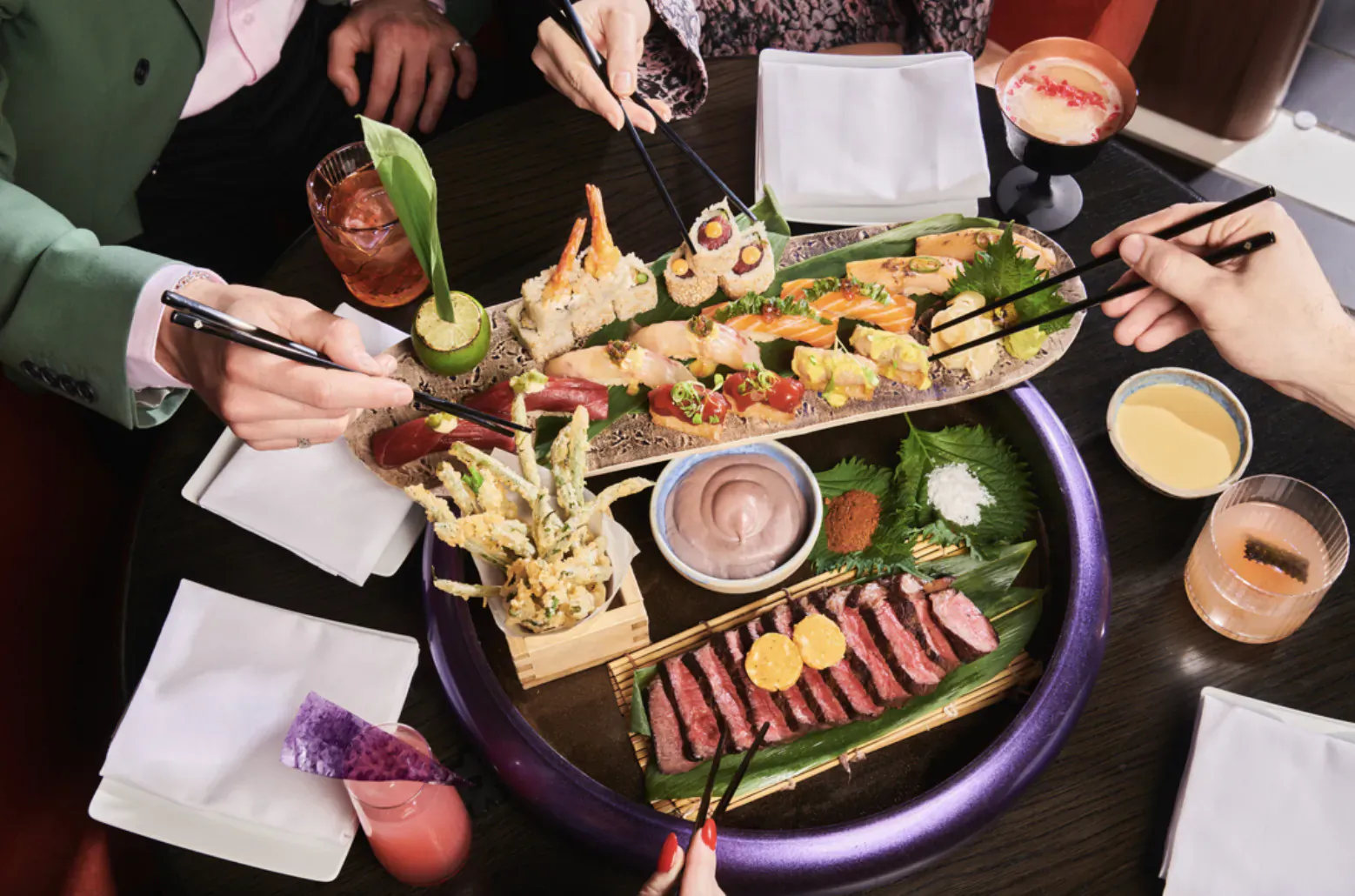 Overhead view of diverse sushi platter with chopsticks held by hands, on wooden table with cocktails.