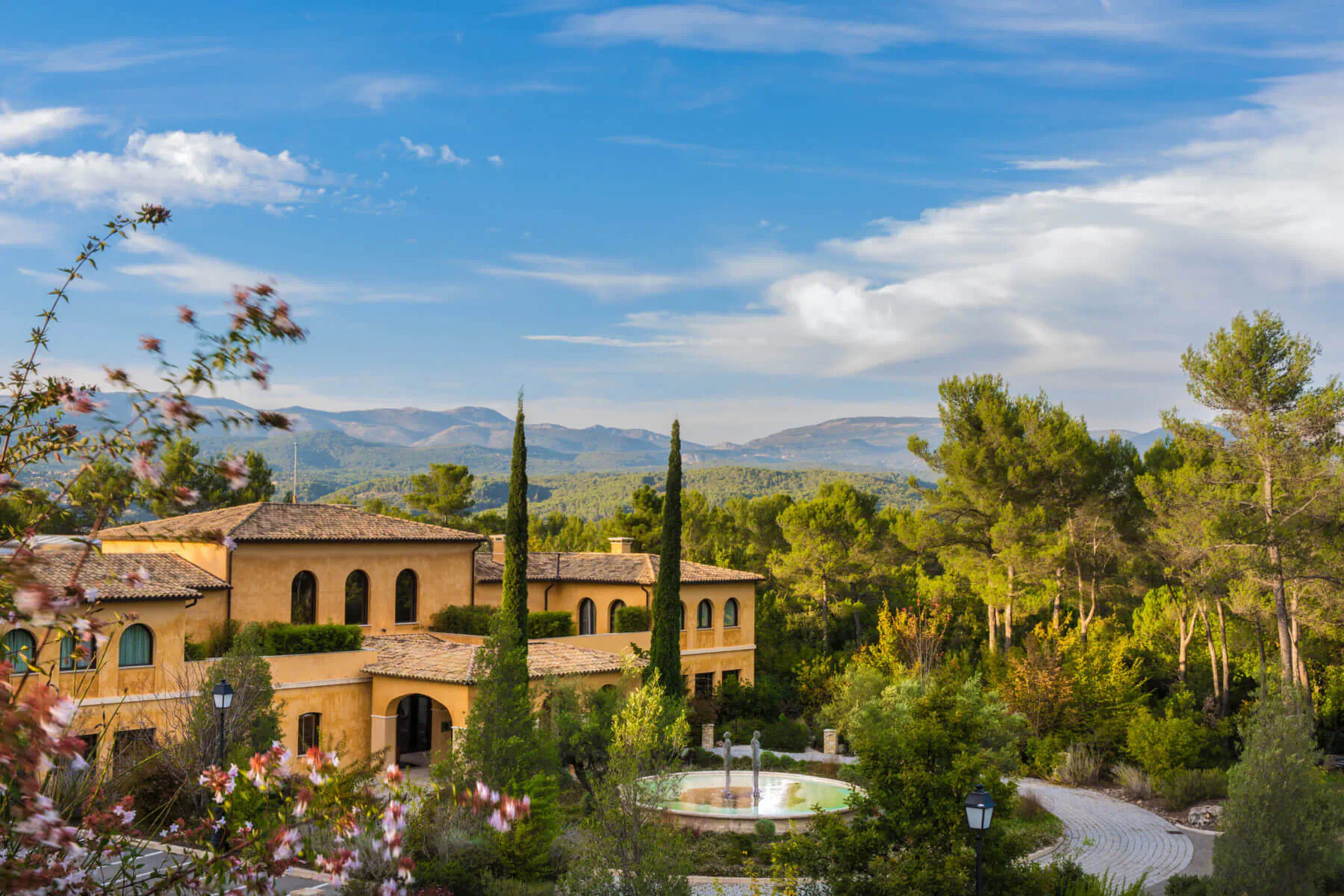 Aerial view of 5-Star Terre Blanche Hotel Spa Golf Resort, yellow villa amid pine forests and mountains in Provence, with fountain and gardens.