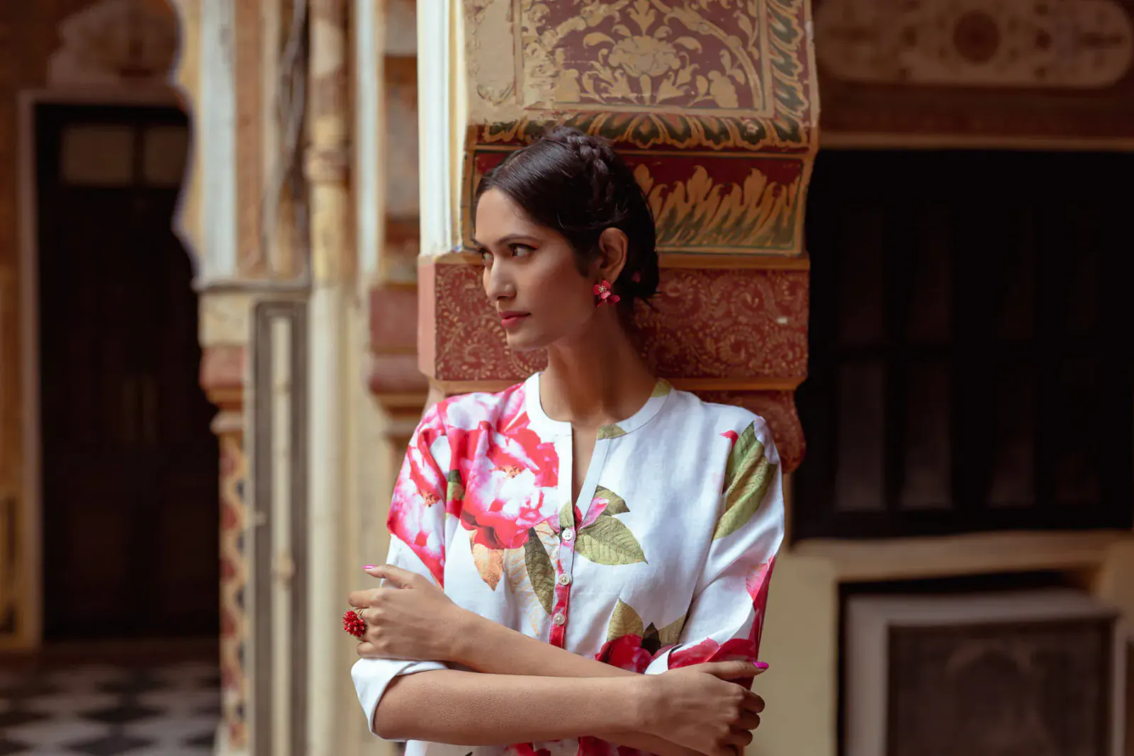 Indian woman in floral white kurta with pink earrings, arms crossed, leaning against ornate pillar in courtyard