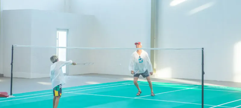 Two men playing badminton on green court in indoor gym at Hideaway Beach Resort, one smashing shuttlecock over net.
