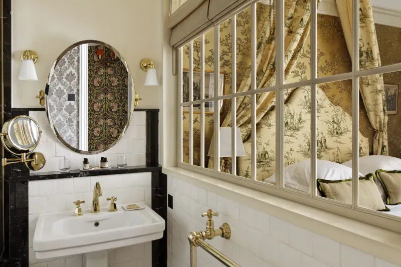 Elegant bathroom in Relais Christine Paris with gold sink, oval mirrors, and floral bedroom view through French windows.