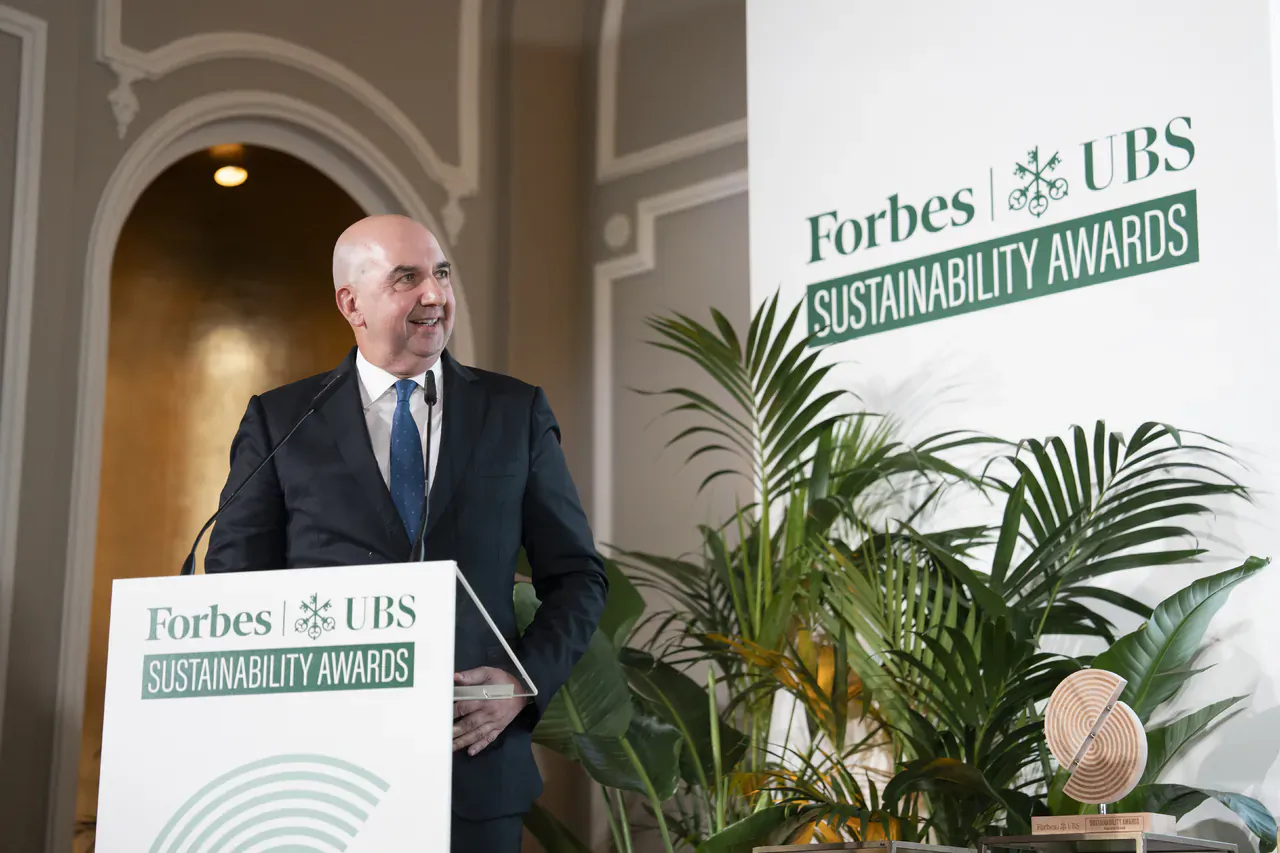 Ricardo Fisas, bald man in suit, speaking at Forbes x UBS Sustainability Awards podium with plants and backdrop.