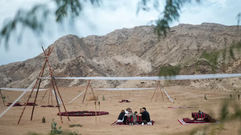 People in traditional attire picnic on red rugs amid teepees and nets in desert with mountains, luxury Zerzura setup.