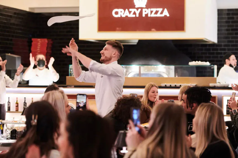 Man in white shirt tosses pizza dough in front of 'CRAZY PIZZA' sign, cheering crowd at National Pizza Day celebration.