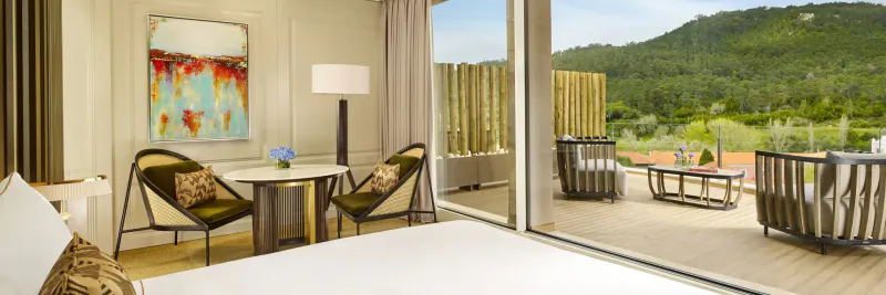 Luxurious hotel room with king bed, lounge chairs, modern art, and sliding doors to terrace overlooking green hills and golf course.