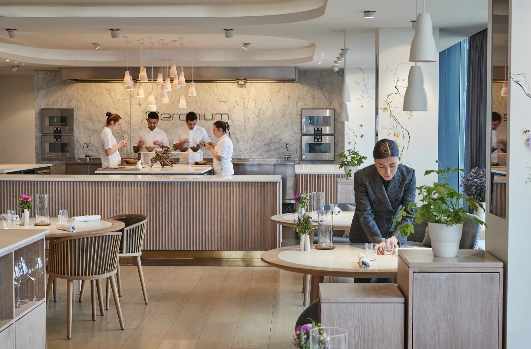 Geranium Restaurant: chefs preparing dishes in modern kitchen, woman working at wooden table with plants.
