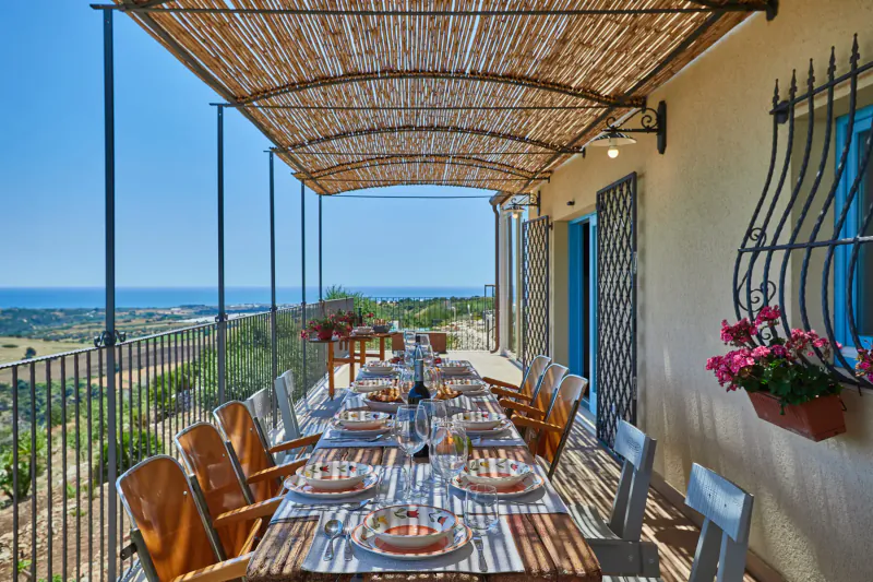 Luxury Sicilian villa terrace with long set dining table, bamboo pergola, pink flowers, and panoramic sea views.