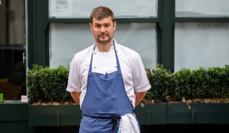 Chef in white shirt and blue apron stands outside 1771 Restaurant on Holbein Place, with green shrubs and windows behind.