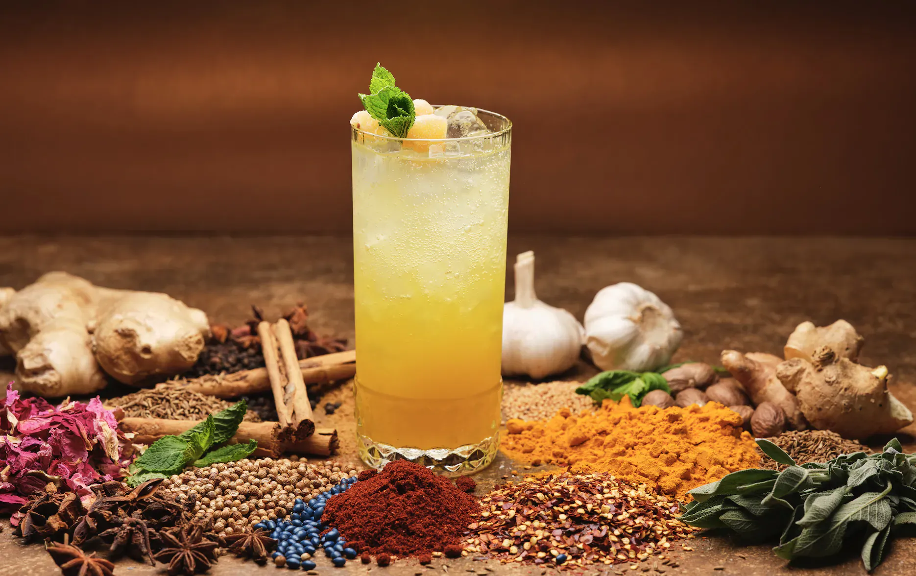 Tall yellow cocktail garnished with lime and mint, surrounded by ginger, garlic, spices, herbs on wooden table