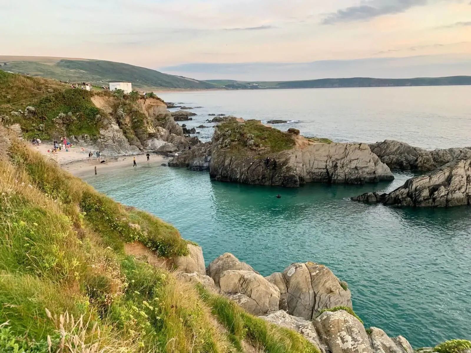 Woolacombe Bay in North Devon: turquoise cove beach with cliffs, rocks, sea, and people at sunset.