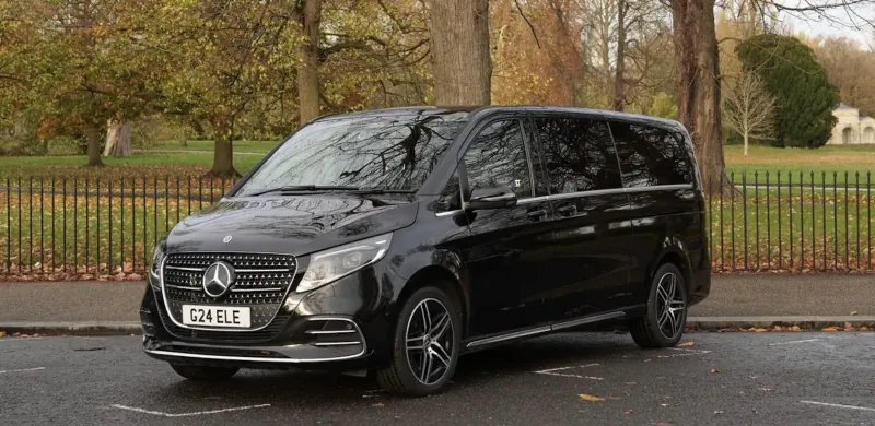 Black Mercedes-Benz V-Class luxury van parked on wet street in autumn park, Mercedes plate visible.